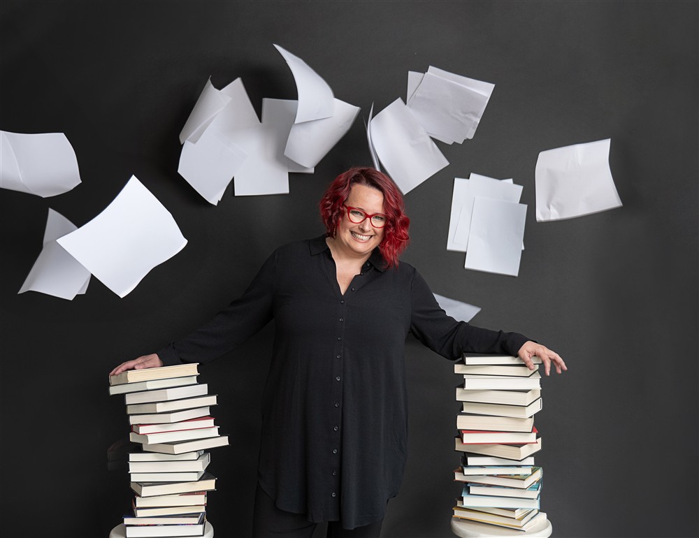 Jocelyn Lindsay smiling between stacks of books with pages flying overhead
